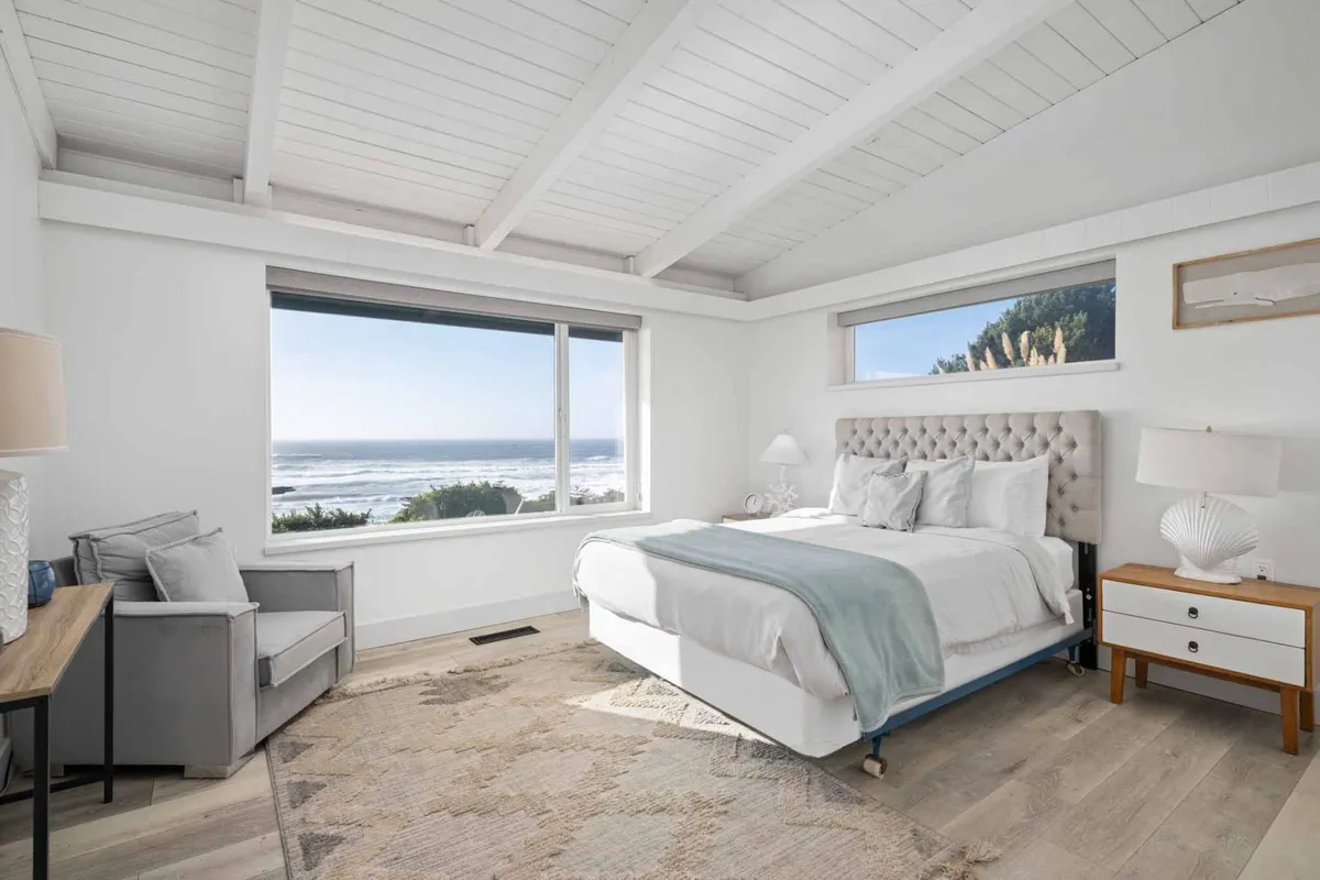 A spacious, sunlit bedroom with a plush bed facing a large window offering stunning ocean views. An armchair sits in the corner next to a nightstand with a lamp, creating a serene spot for relaxation. The room feels airy and restful, with soft tones and coastal accents.