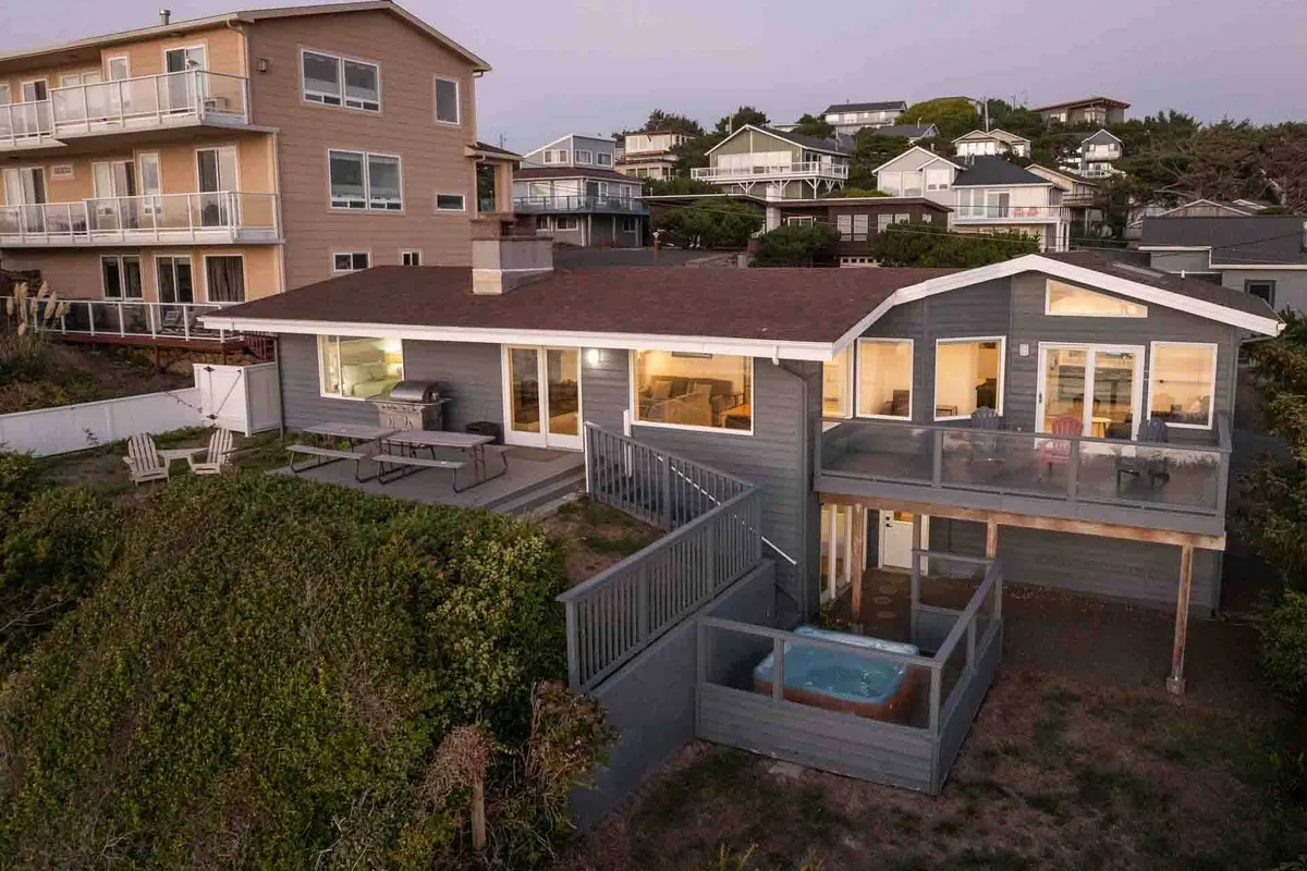 An exterior view featuring a hot tub and outdoor seating area overlooking the ocean, with large windows reflecting the landscape. The serene setup suggests relaxation while immersed in natural beauty, ideal for quiet evenings or stargazing.