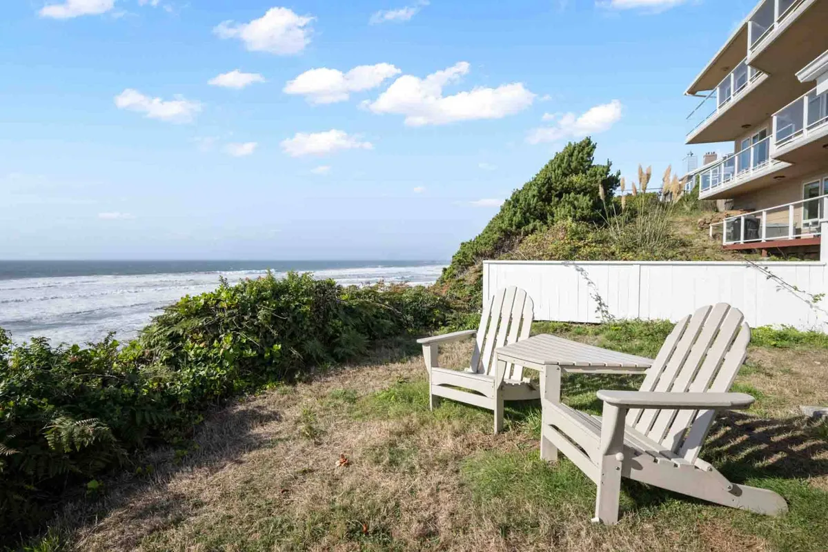 This image displays a set of Adirondack chairs facing the ocean with lush landscape greenery surrounding the area. The tranquil setting invites guests to relax, enjoy the ocean view, and feel the sea breeze, clearly showcasing an exterior living space connected to the coastal environment.