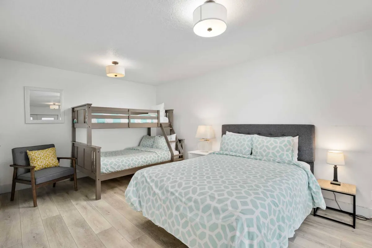 A comfortable bedroom featuring both a bunk bed and a queen bed, complemented by an armchair and a nightstand with ambient lighting. The room is spacious and inviting, making it ideal for families or groups with children.