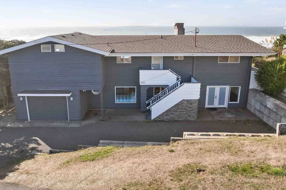 This exterior image features the modern architectural design of Moonstruck Beach House, set against expansive ocean views. The staircase leads to the inviting entrance, and the porch offers a tranquil spot to soak in the coastal surroundings. The ocean is visible in the background, establishing a strong sense of place and proximity to the beach.