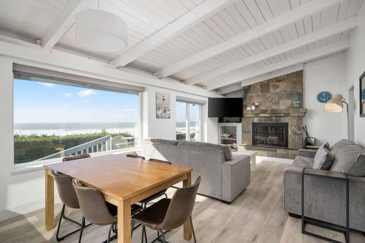 The hero image captures a spacious, inviting living and dining area with expansive windows framing sweeping ocean views. The stone fireplace adds a cozy focal point, while a large dining table promises memorable shared meals. Natural light floods the space, highlighting comfortable furnishings and drawing the eye to the dramatic Pacific backdrop.