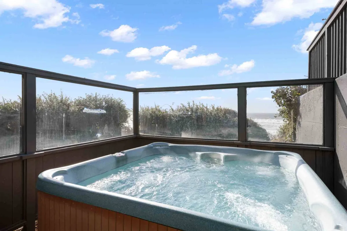 A cliff-top hot tub positioned with a panoramic ocean view, surrounded by natural greenery. The hot tub area is partially shielded, allowing for relaxation while taking in the expansive sky and sea. The setting suggests tranquil moments at sunset or under the stars, with the soothing sound of waves in the background.