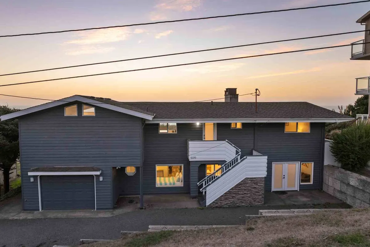 The image shows the double-story façade of the Moonstruck Beach House, with a staircase leading to an inviting entrance and large windows glowing from interior lighting. The scene is serene and welcoming, suggesting a friendly, family-oriented retreat.