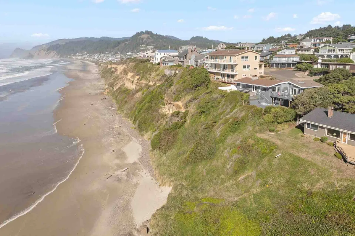 This image captures cliffside houses overlooking a broad sandy beach and rolling ocean waves, with distant mountains on the horizon. The composition speaks to the region's natural beauty and the abundance of coastal adventures available, from tide pool exploration to catching sunsets from scenic vantage points.