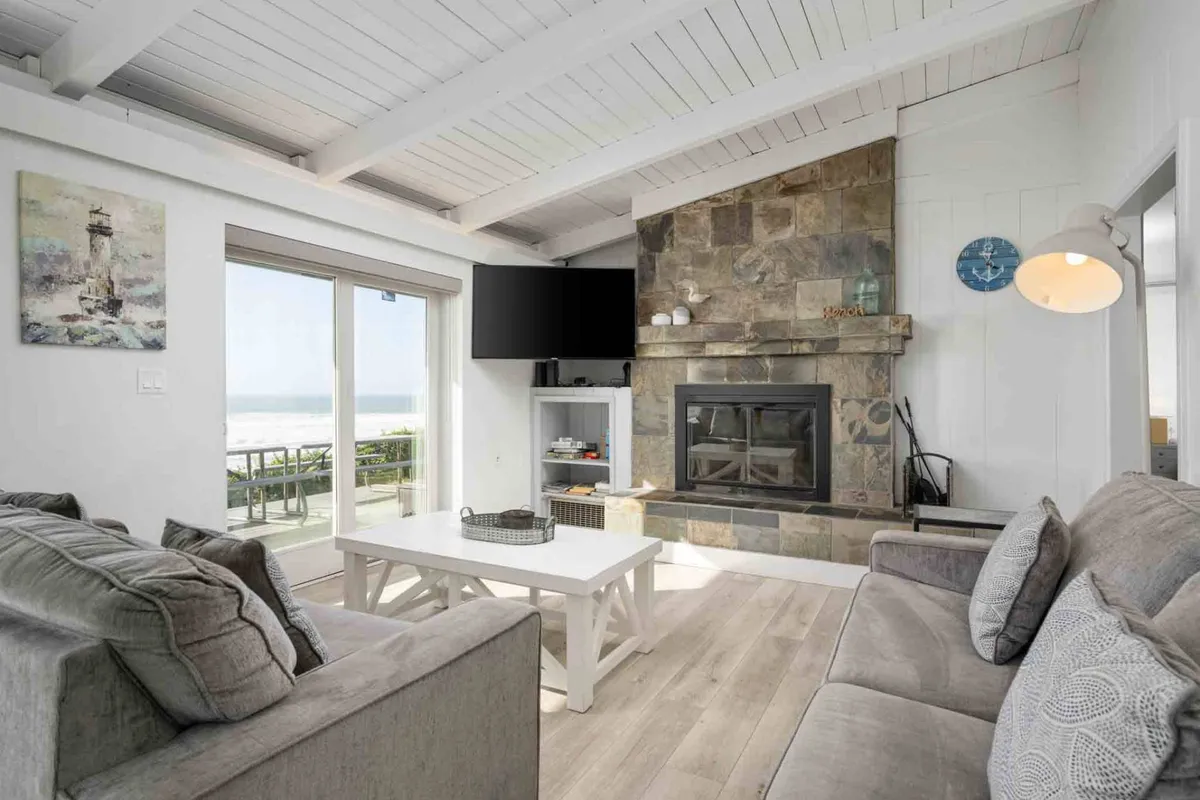 A cozy stone fireplace anchors the living room, surrounded by a comfortable sofa and coffee table, with sliding glass doors that frame the ocean beyond. The room is flooded with natural light, creating an inviting space for relaxation, storytelling, or enjoying the view.
