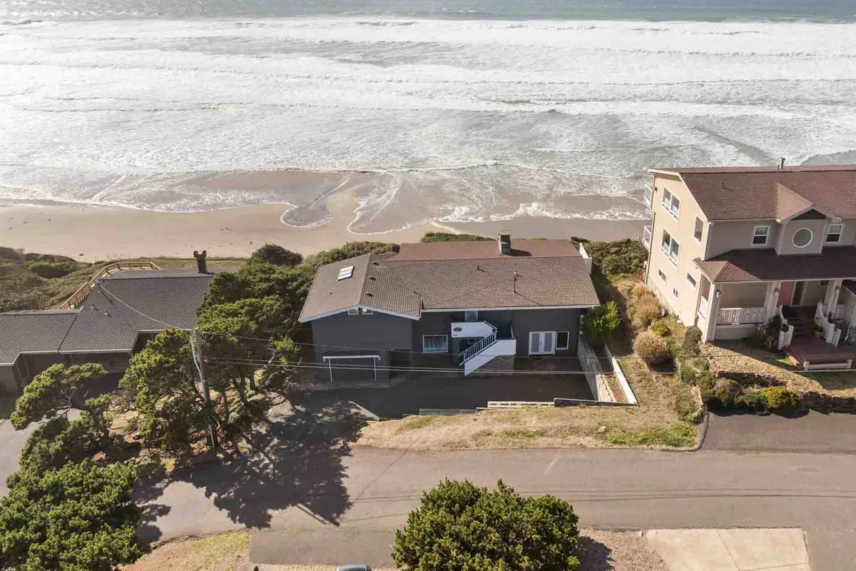 An inviting beachfront view showing the waves crashing on the sand, lush greenery around the property, and neighboring homes dotting the coastline. The image evokes an active beach environment ideal for sandcastle building, beachcombing, and family gatherings.