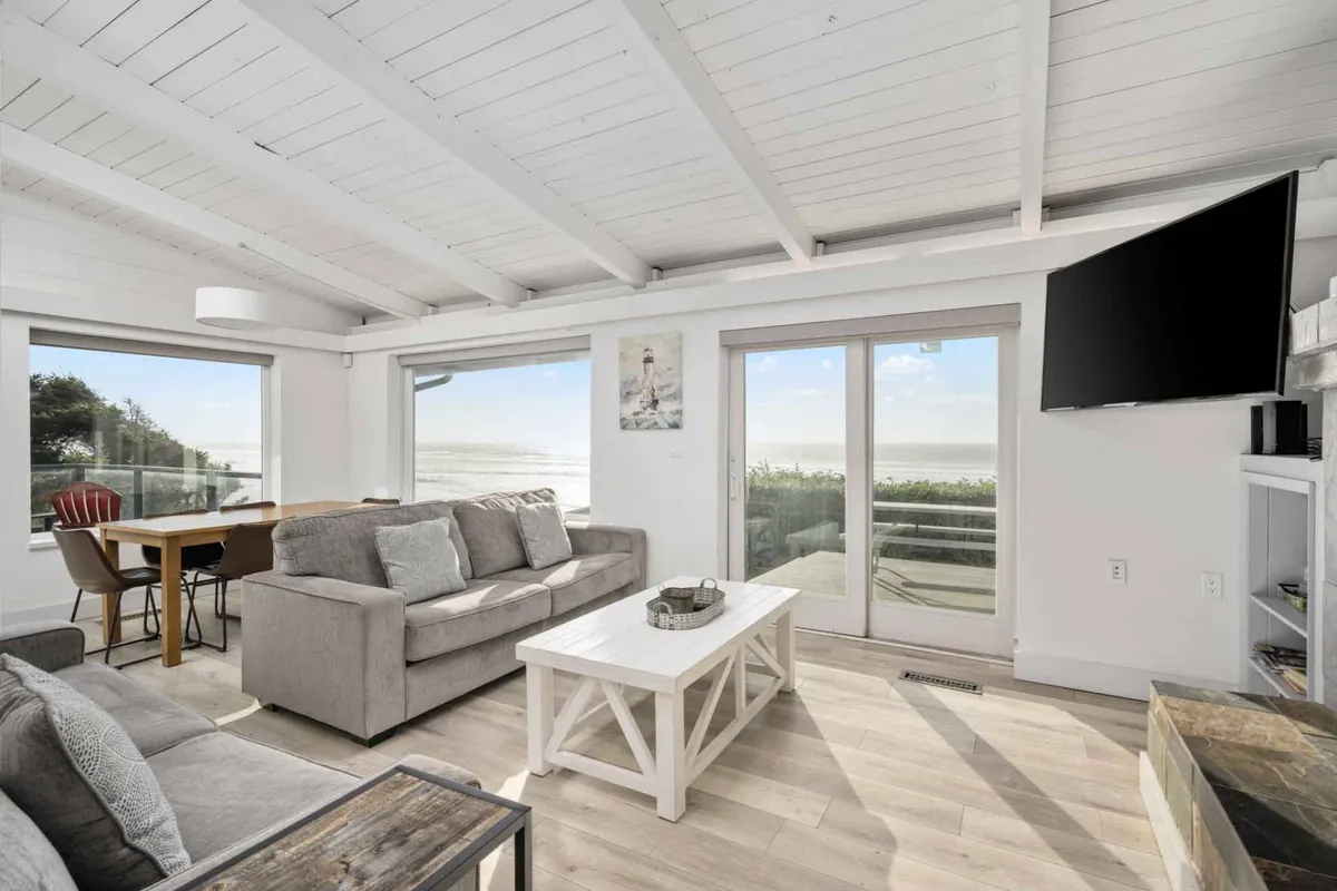 A spacious, open-plan living and dining area filled with light from large windows that showcase an expansive ocean view. Comfortable seating and a dining table are arranged to encourage socializing, relaxation, and shared stories. The setup is ideal for group gatherings, playing games, or simply enjoying the scenery together.